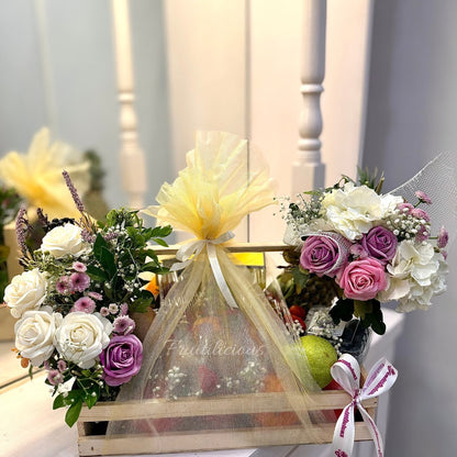 A fresh fruit basket containing a variety of fruits and flowers, neatly arranged in a wooden basket with a ribbon on the side.